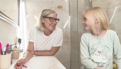 Woman helping her daughter brush her teeth in front of a running faucet