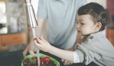 Woman helping her daughter brush her teeth in front of a running faucet
